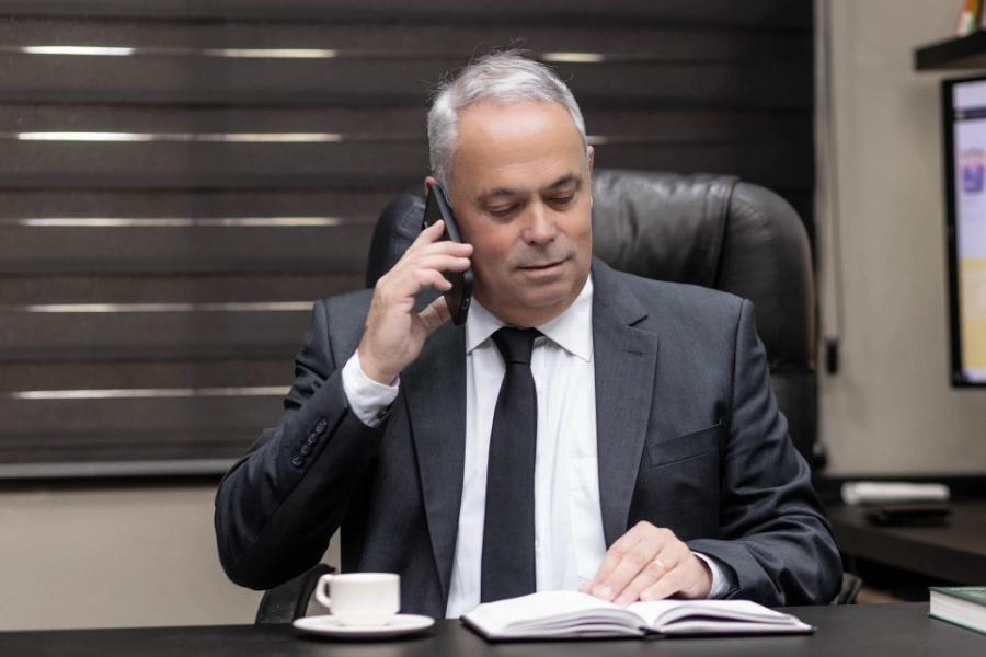 Carlos Gomes no escritório, em ligação telefônica, com uma xícara e livro abertos sobre a mesa.
